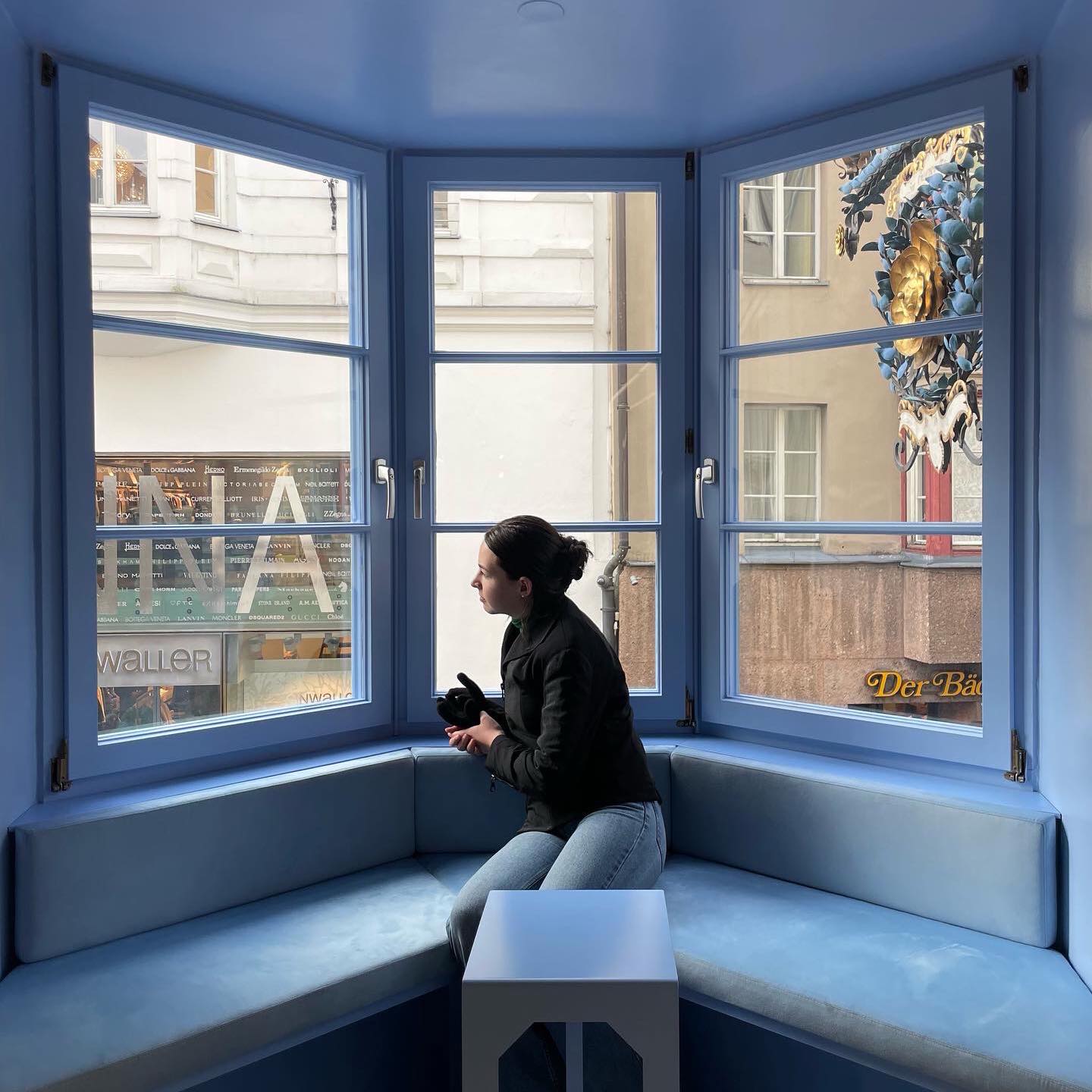 Page looking out a window inside Swarovski Innsbruck. She is sitting by a light blue bay window, looking out at people in the world. Her brunette hair is in a clip, with her body directed towards the left window.