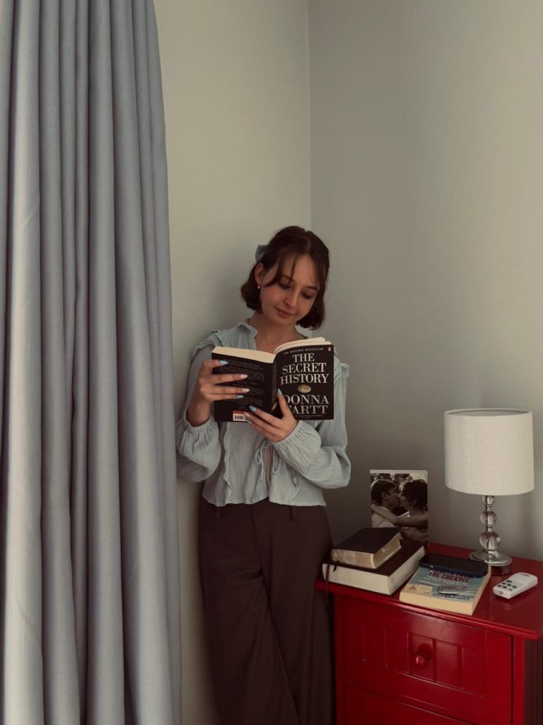 Page leaning on a wall, reading a book called the secret history.